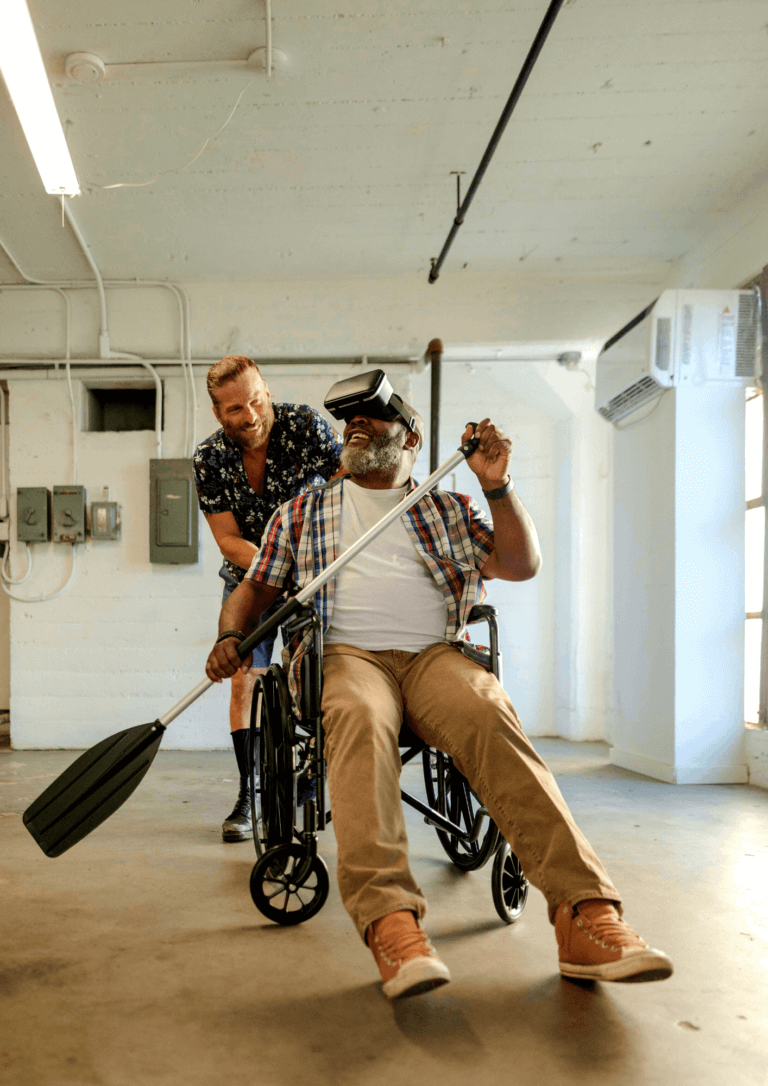 A carer pushing a Man in a wheelchair wearing VR or a virtual reality headset whilst holding a boating ore having fun in a warehouse